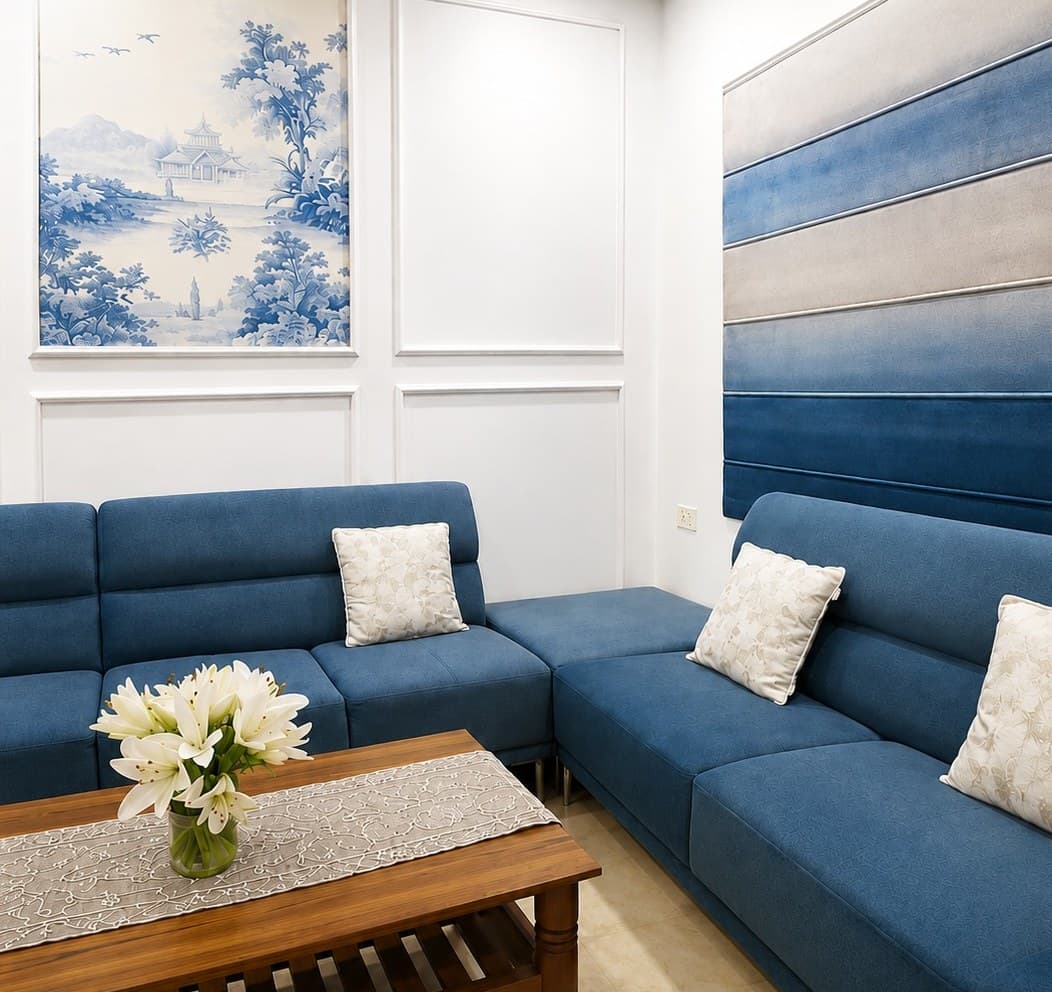 A living room on Road No. 12, Banjara Hills — a blue sectional sofa with cream cushions, white panelled walls, a blue-and-white landscape painting, and a wall of upholstered panels in bands of blue and grey.