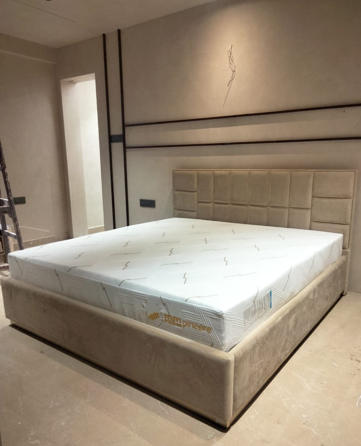 A second bedroom in the same flat during the fit-out, with a wide square-grid bouclé headboard on a beige upholstered bed base and an MM Foam Pincore mattress still in its wrapper on top, a stepladder and unfinished walls either side.