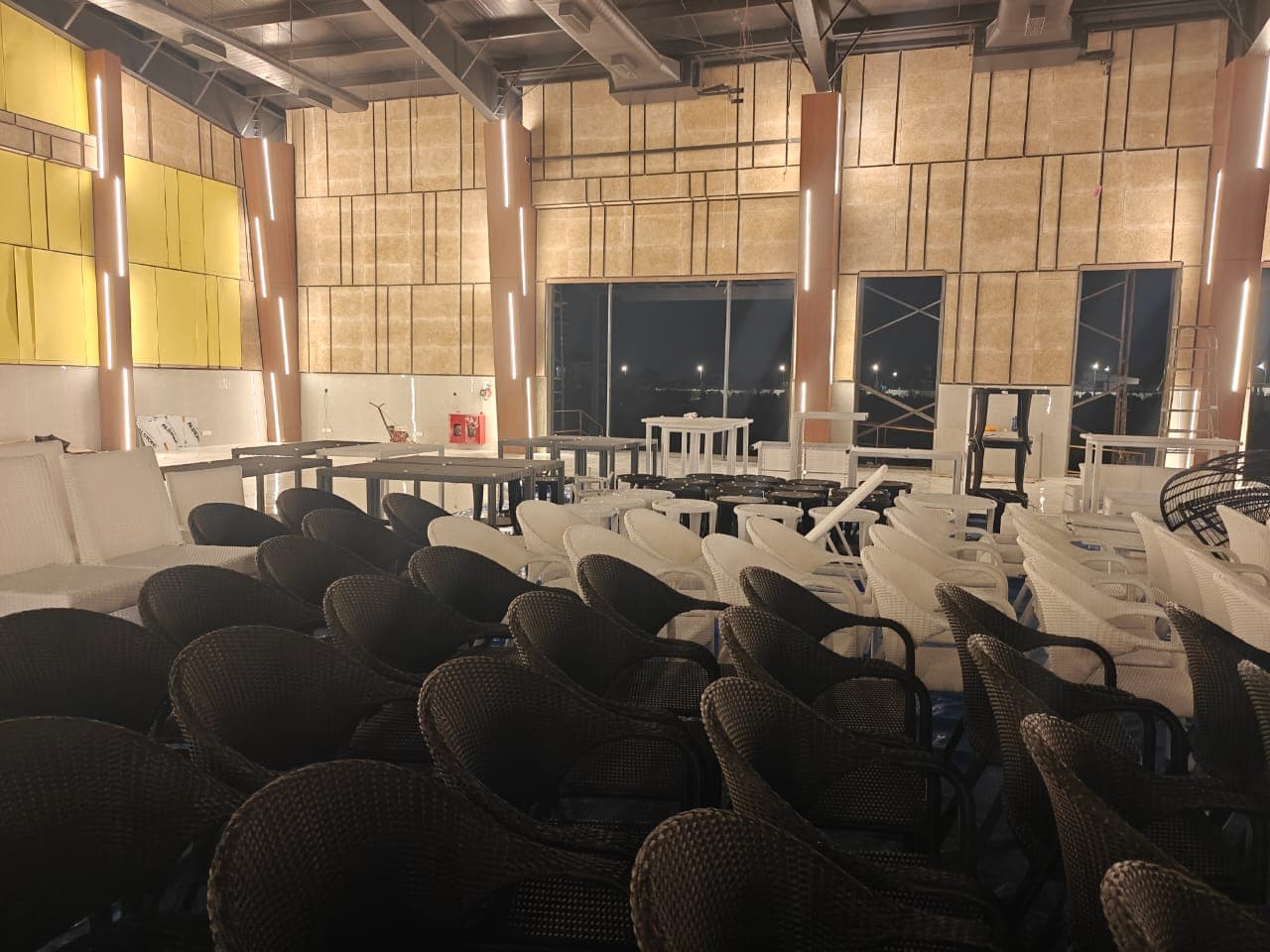Woven chairs and tables laid out across a tall hall at Lore Resorts and Conventions at night during fit-out, lit by vertical strip lights on the columns.