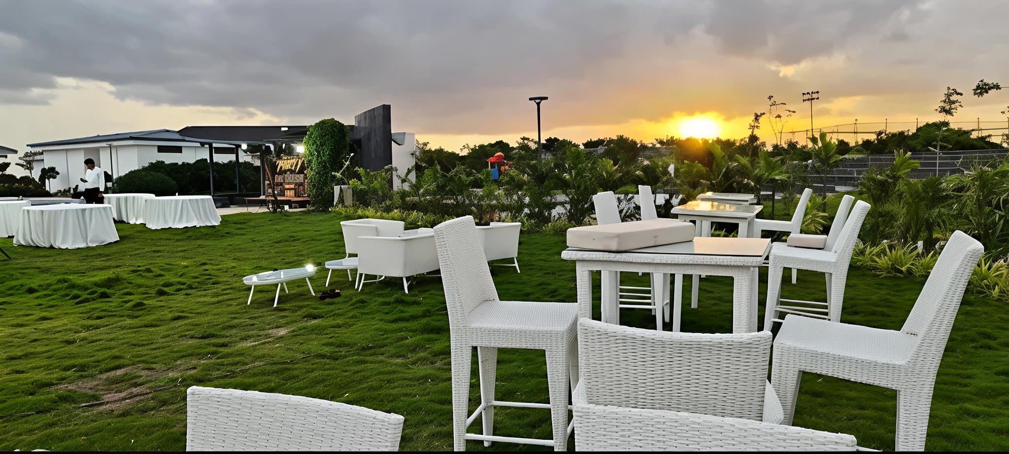 Woven outdoor furniture at Lore Resorts and Conventions laid out on the lawn at sunset — bar-height tables and chairs, low seating and cushioned benches, with the pavilion behind and the sun going down through the planting.
