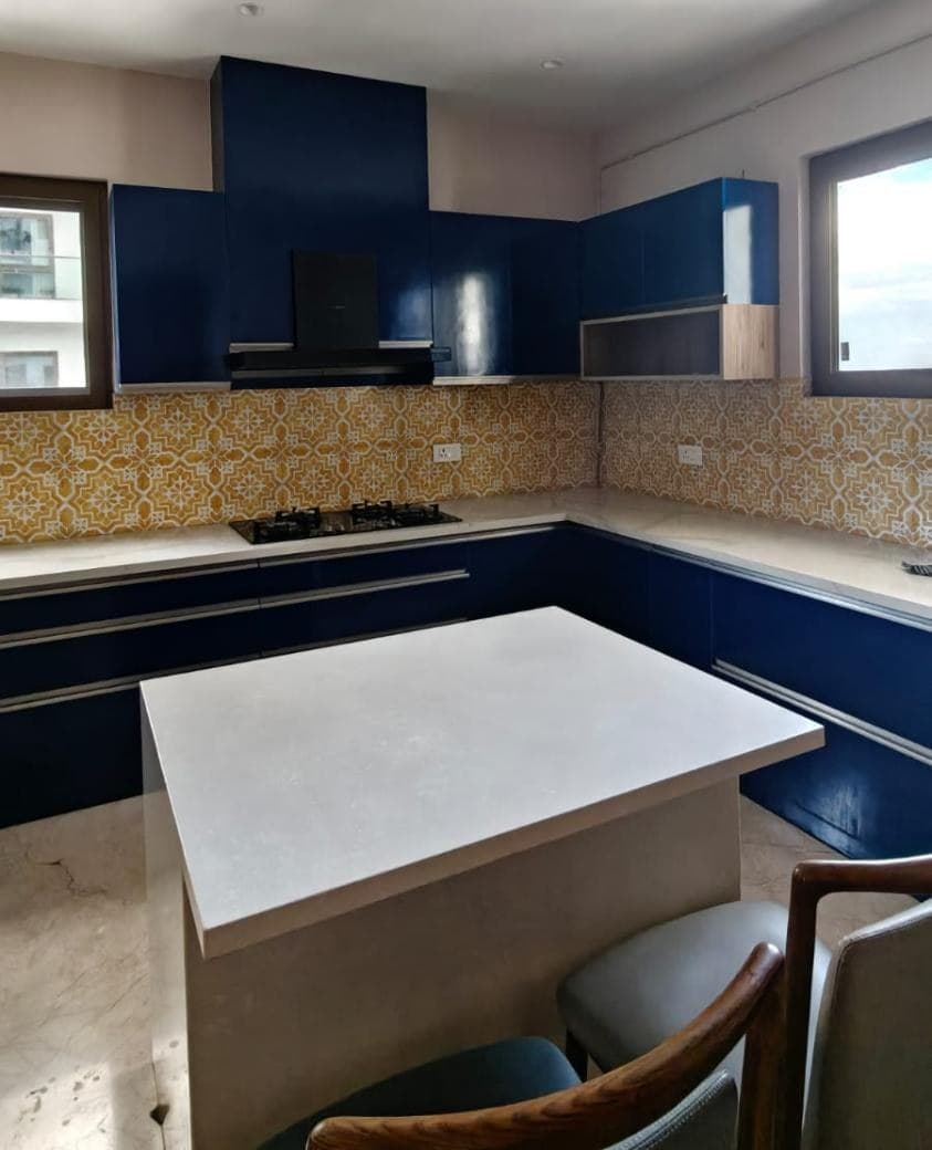 A finished L-shaped kitchen in Hyderabad — gloss navy units on two walls, a black glass chimney boxed into the run, a patterned yellow splashback, and a free-standing island with a light stone top and chairs pulled up to it