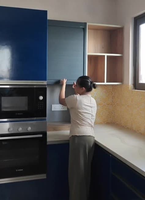 Somebody lifting the tambour rolling shutter of an appliance garage in the same kitchen, with the built-in microwave and oven in the tall unit beside it and open shelves to the right