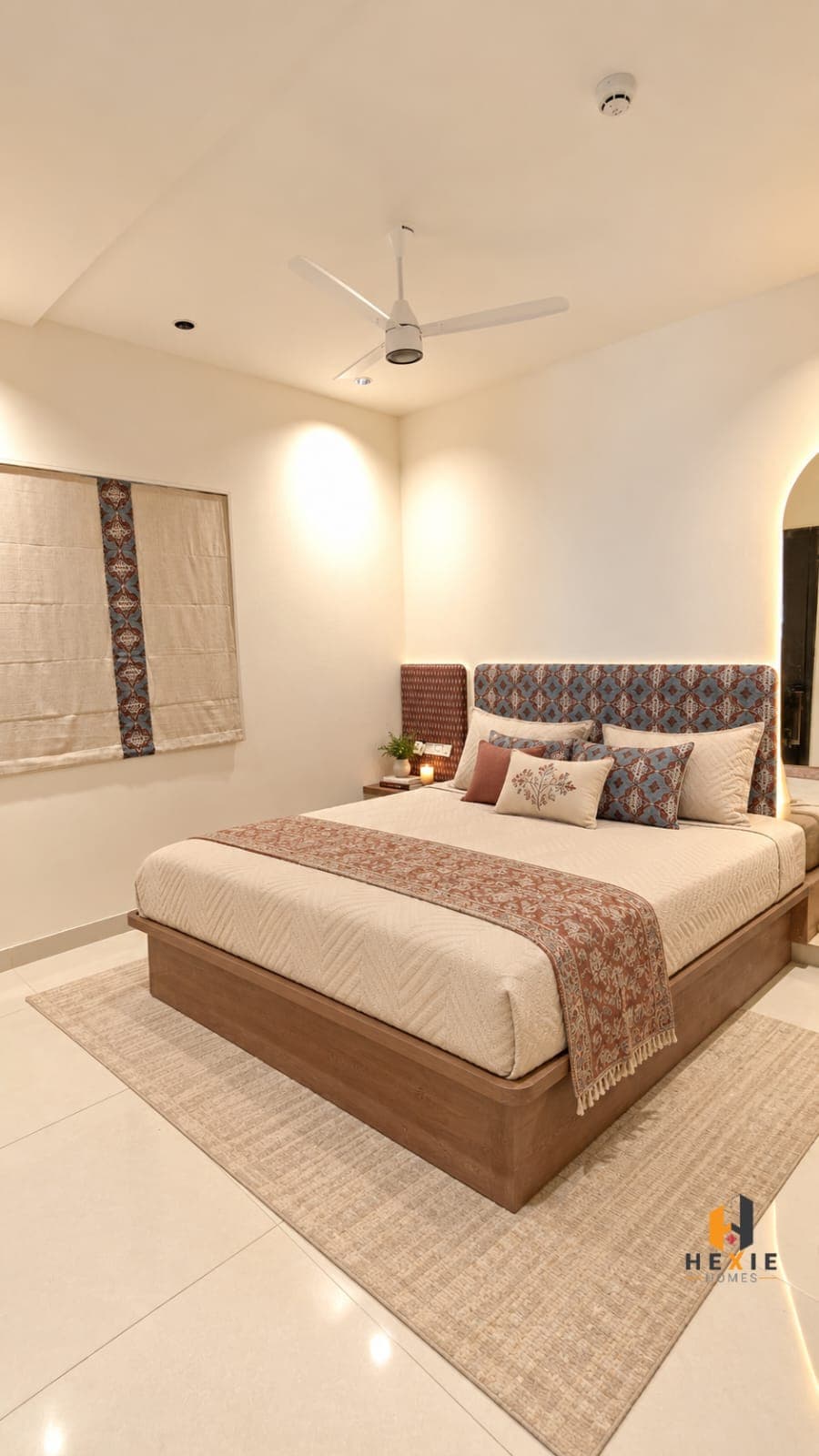 The second bedroom, with a headboard upholstered in a blue and rust ikat pattern and lit from behind, a timber bed frame, a rust bed runner, and a natural-cloth Roman blind with a printed border band down one side.
