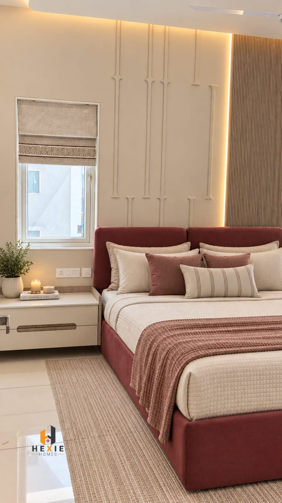 A bedroom with a maroon velvet upholstered headboard against a cream moulded wall lit from behind, cream and rust bedding, and a Roman blind in textured neutral cloth carrying a fret-pattern border band across it.