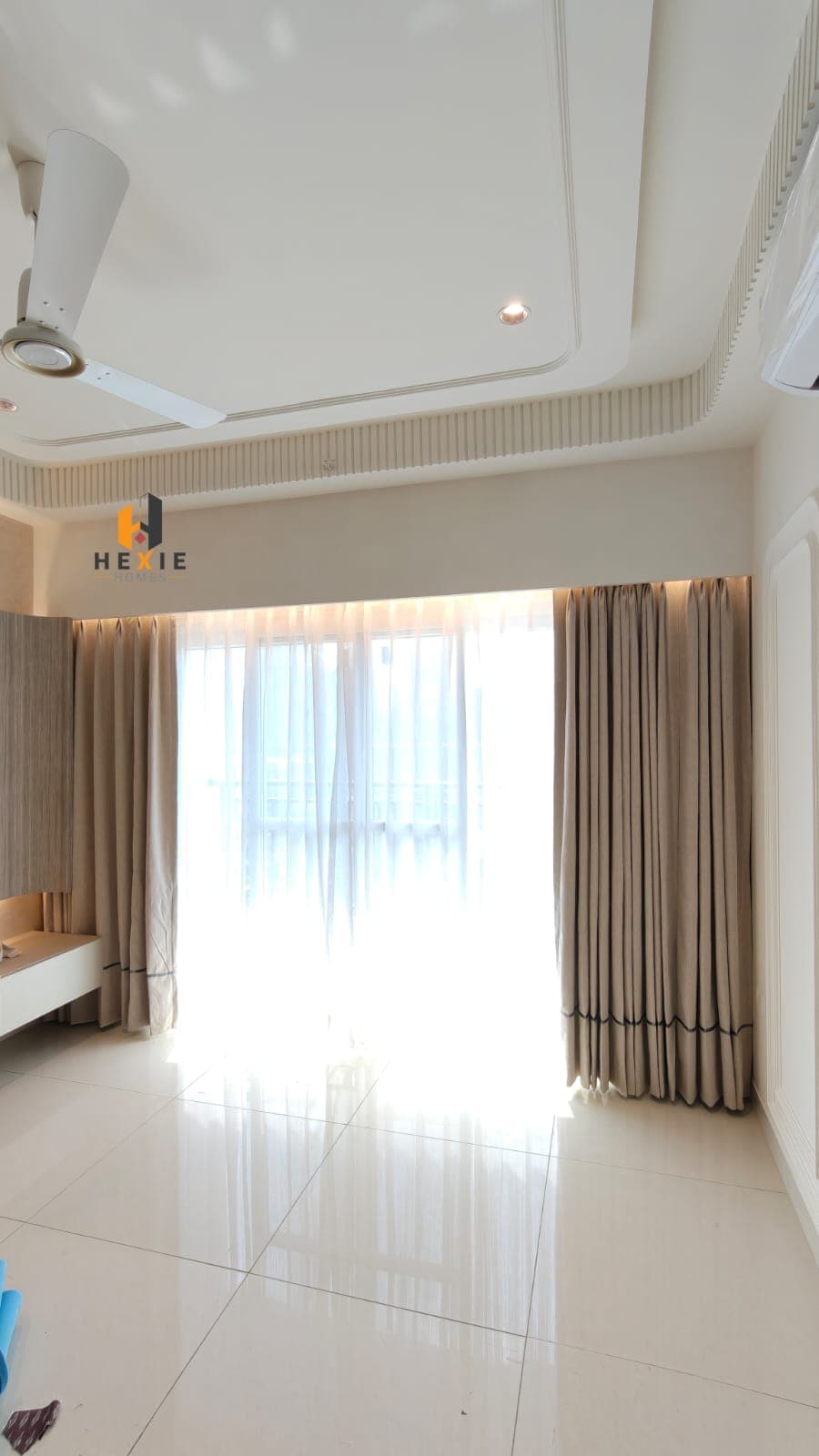 A full-height window at Aparna Zenon with a white sheer drawn across it and a pair of stone-coloured curtains either side, each carrying a dark contrast border up the leading edge and along the hem, hung from a recess in the false ceiling with a warm light above them.