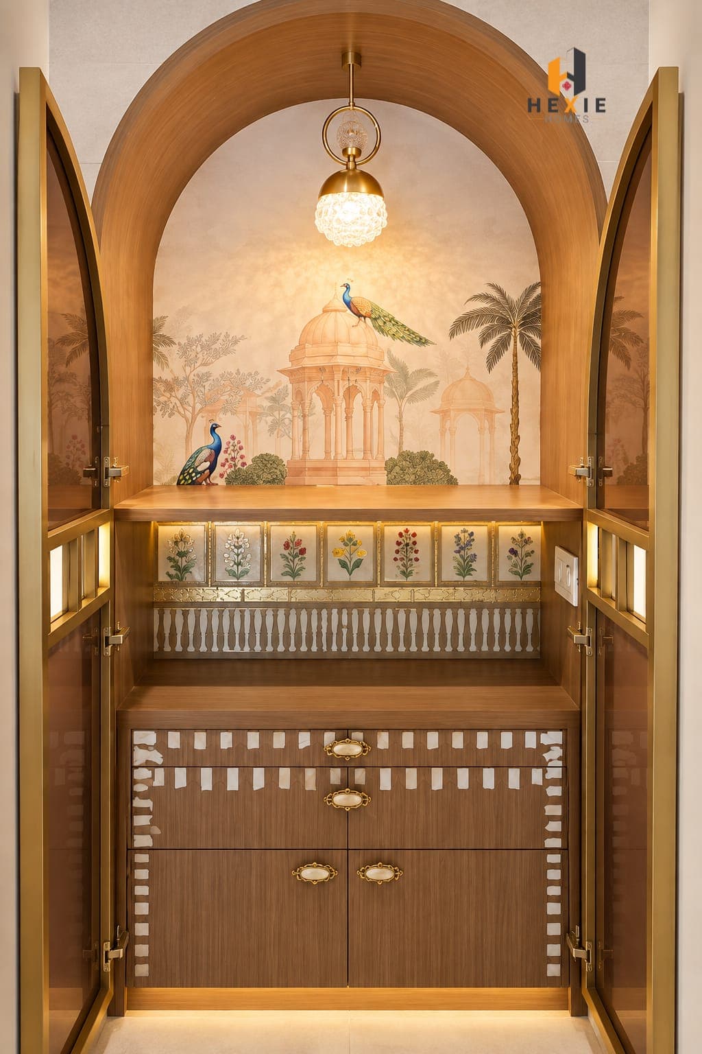 The mandir — an arched timber unit behind brass-framed glass doors, with a peacock and chhatri mural papered across the back wall, seven small botanical panels lit along the shelf, and a brass and glass pendant hanging in the arch.