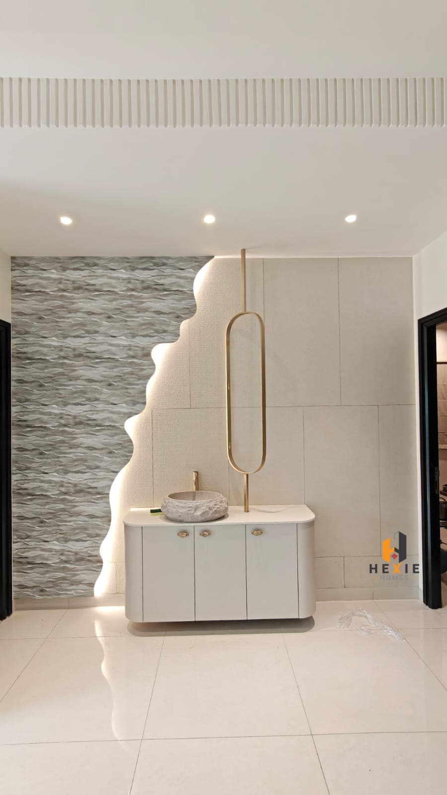 A washbasin against a two-part wall — a grey three-dimensional ribbed panel meeting a cream textured wallpaper on a scalloped edge that is lit from behind — with a stone vessel basin on a curved white vanity and a tall brass mirror above it.